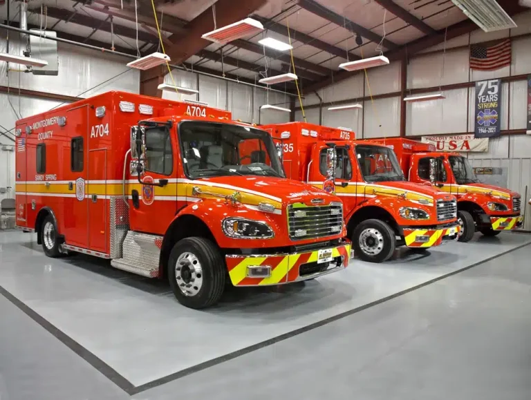 Three bright red emergency vehicles with yellow stripes, including Ambulance 704 Sandy Spring VFD, are parked inside a clean, well-lit garage. Overhead lights reflect off the vehicles. A banner honoring 75 years of service hangs on the back wall.