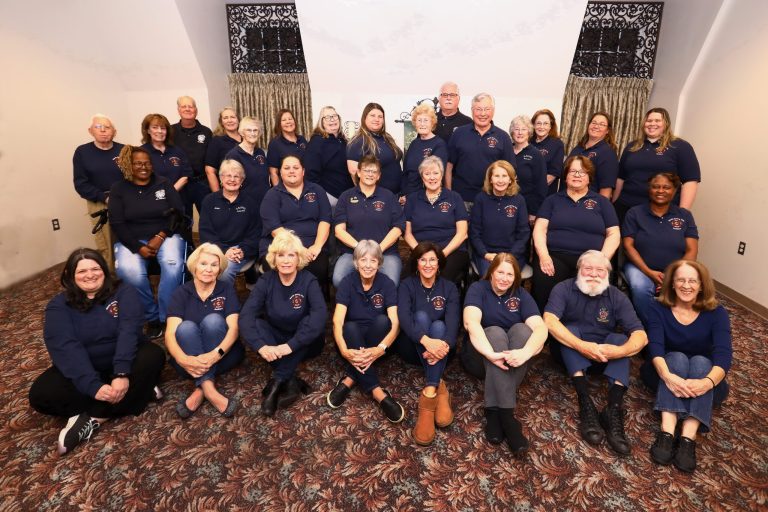 A group of men and women in matching navy shirts pose together indoors, sitting and standing in rows on a patterned carpet in front of light-colored walls and curtains—a testament to SSVFD history and the spirit of the Montgomery County fire department.