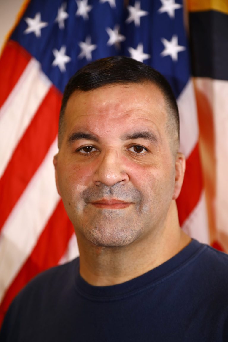 Steve Hamatto, wearing a blue shirt, stands in front of a blurred American flag background, looking at the camera with a neutral expression. As a member of the Sandy Spring Volunteer Fire Department, his dedication is reflected in his poised demeanor.