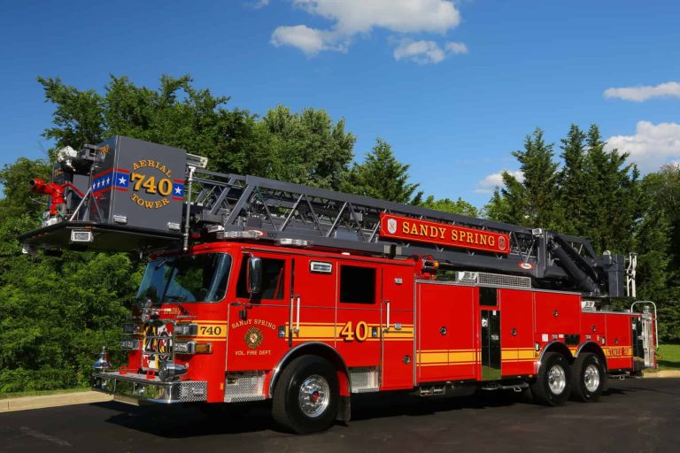 A bright red Aerial Tower 704 Sandy Spring VFD fire truck with an extended ladder is parked on a road, with green trees and a blue sky with scattered clouds in the background.