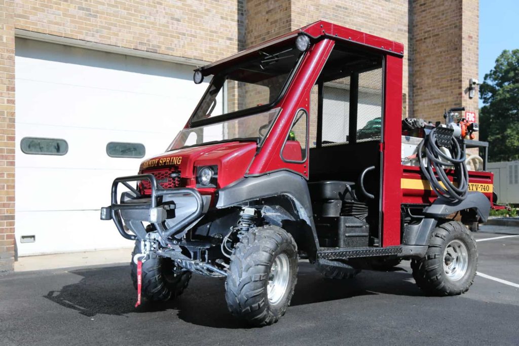 A small red fire utility vehicle labeled BUTV 740 Sandy Spring VFD, equipped with off-road tires, is parked on pavement in front of a brick building with a white garage door. The vehicle has hoses and firefighting equipment attached to its rear.