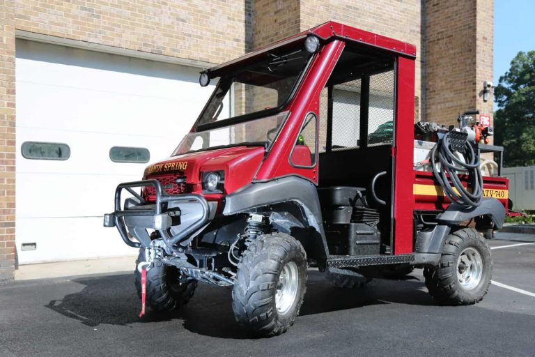 A small red fire utility vehicle labeled BUTV 740 Sandy Spring VFD, equipped with off-road tires, is parked on pavement in front of a brick building with a white garage door. The vehicle has hoses and firefighting equipment attached to its rear.