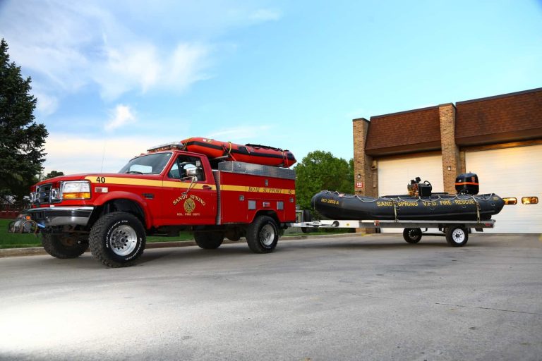 A red rescue truck from the Sandy Spring VFD is parked outside a building, towing Boat 704 Bravo—a black inflatable rescue boat—on a trailer.
