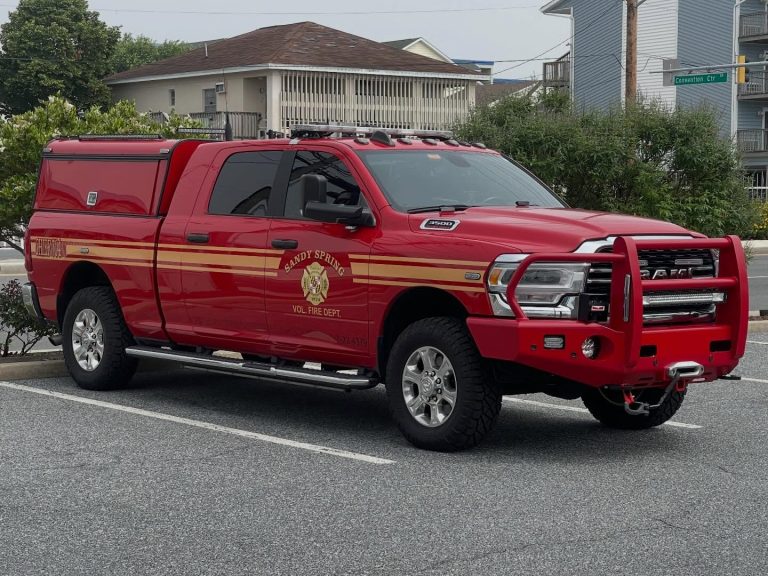 A red pickup truck labeled “Sandy Spring Vol. Fire Dept.” and “Chief 704 Sandy Spring VFD” on the door is parked in a lot near residential buildings. The truck features emergency lights and gold striping.