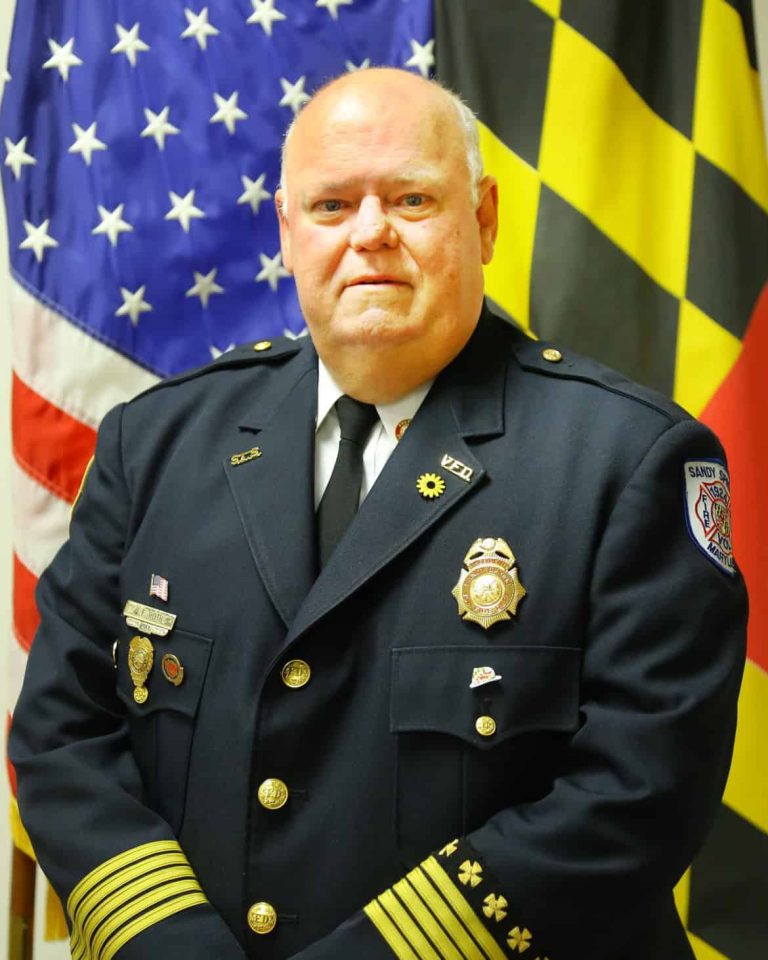 A man in a formal Sandy Spring Volunteer Fire Department uniform stands in front of American and Maryland state flags, looking at the camera. This is Johnie F. Roth, Jr.