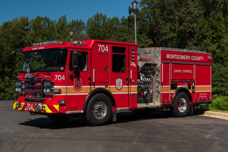 A red Montgomery County fire truck labeled “Sandy Spring” with the number 704, part of Engine 740 Sandy Spring VFD, is parked on asphalt near trees on a sunny day.