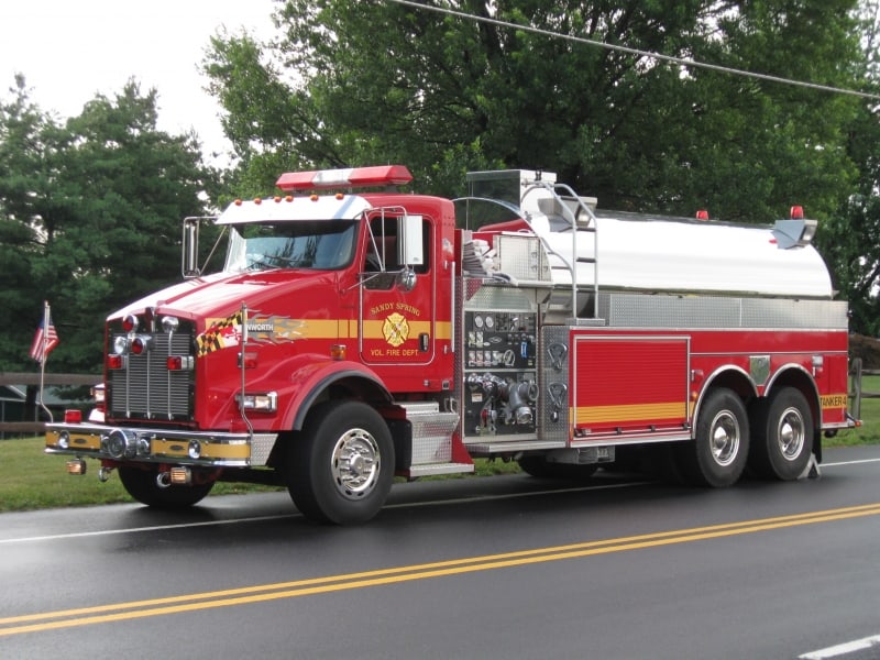 A red fire tanker truck with chrome accents and hoses—identified as Tanker 704 Sandy Spring VFD—is parked on a road near green trees and a wooden fence.