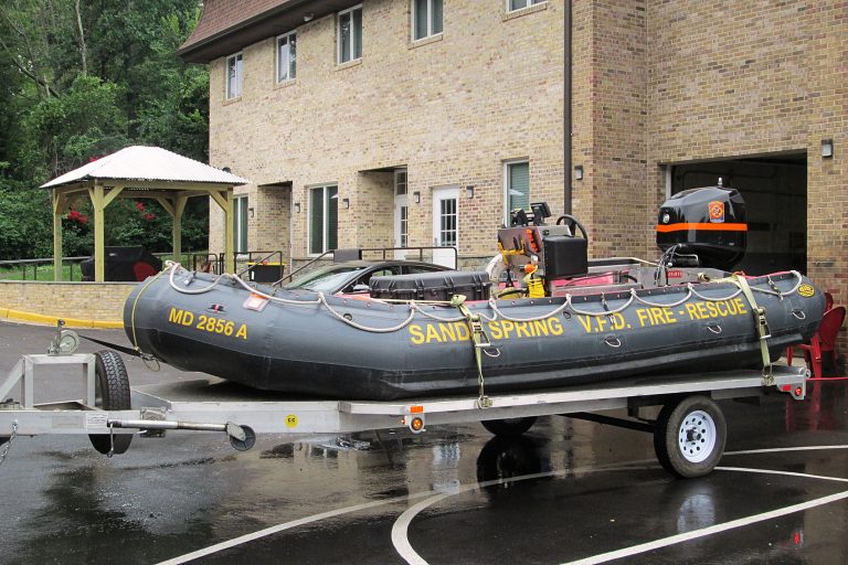 Inflatable rescue boat labeled “Boat 740 Sandy Spring VFD Fire-Rescue” on a trailer in front of a brick fire station building, with wet pavement and a wooden gazebo visible in the background.